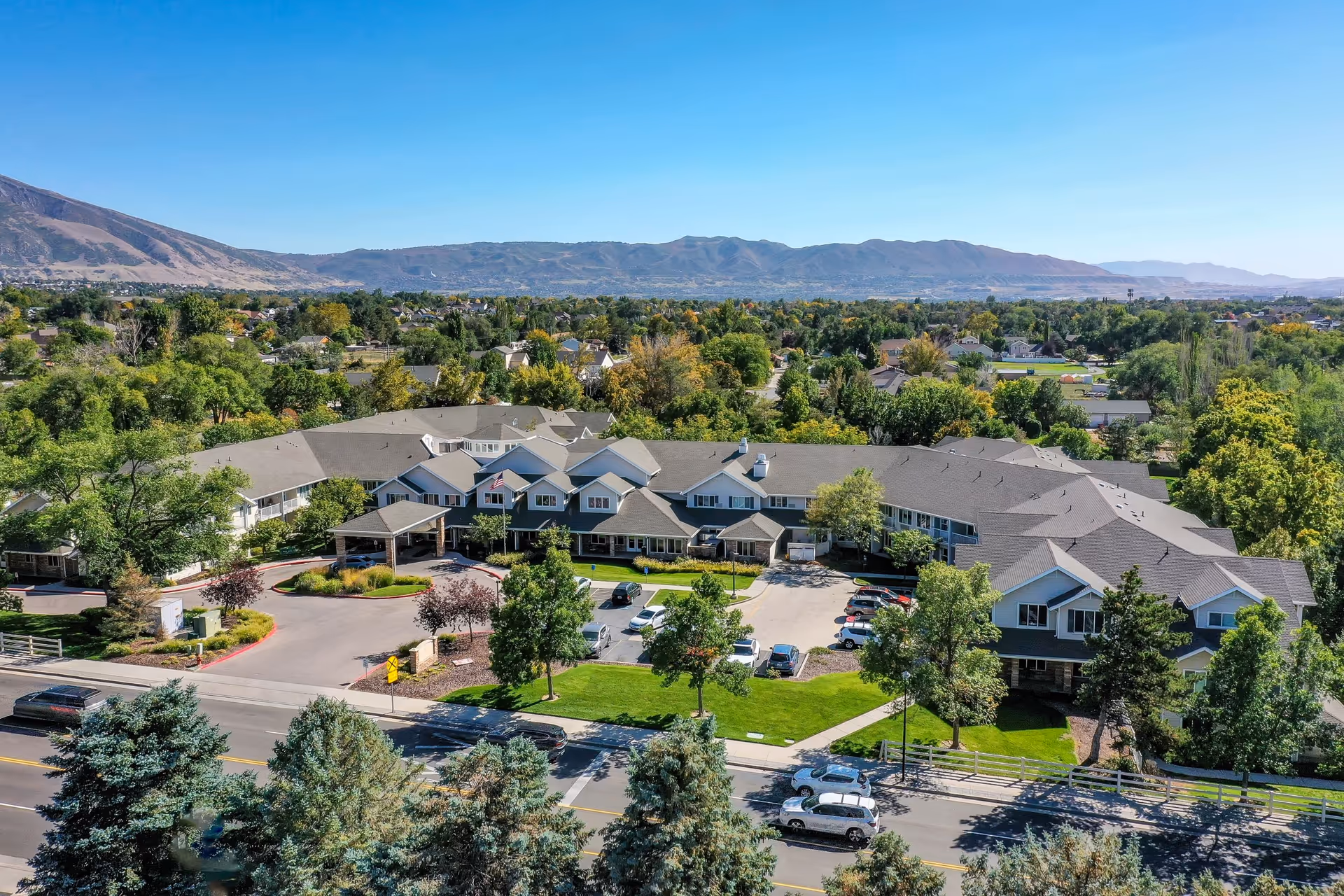 Aerial view of Solstice Senior Living at Sandy, showing a large, multi-wing building surrounded by trees and greenery with a mountain range in the background under a clear blue sky. The facility has a driveway and parking area in front, with several cars parked and well-maintained landscaping.
