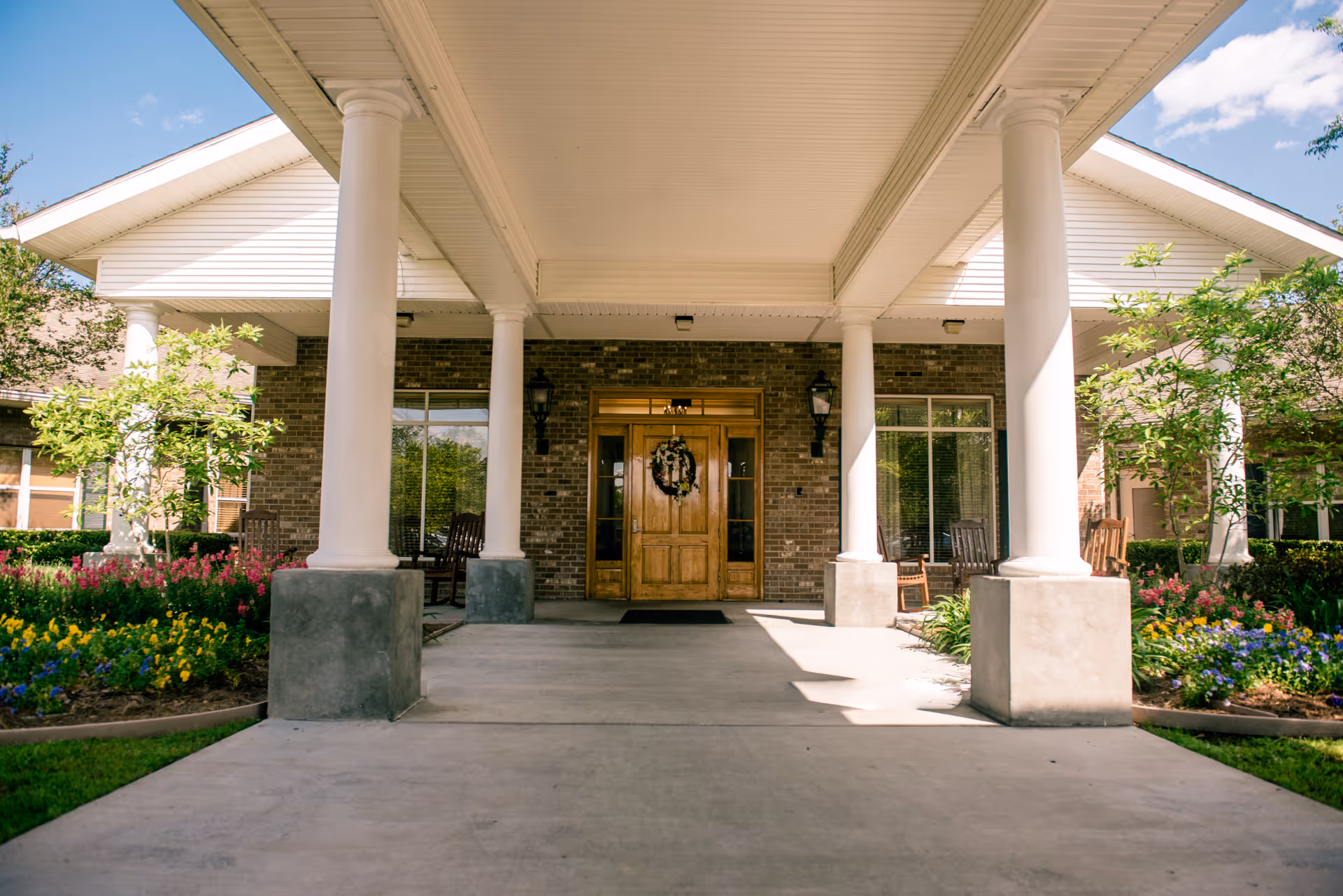Covered entrance of a building with large white columns, wooden double doors with a wreath, and landscaped flower beds.
