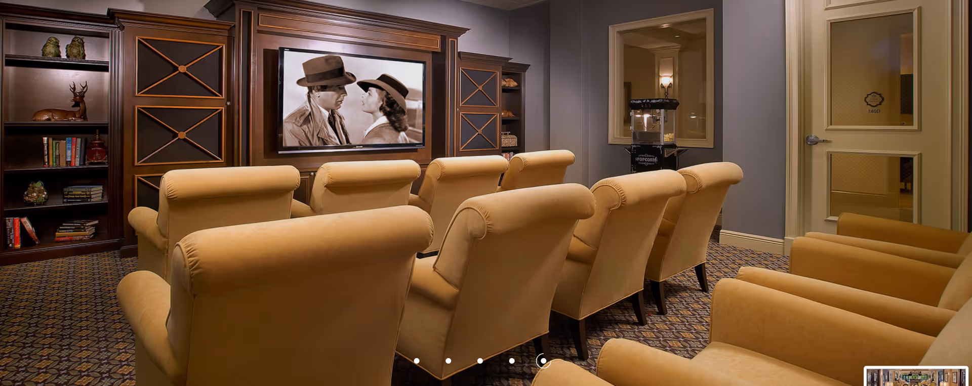 A cozy movie theater room with two rows of beige upholstered armchairs facing a large flat-screen TV mounted on a wooden entertainment center. The TV displays a black-and-white movie scene featuring a man and a woman wearing hats. To the right, there is a popcorn machine next to a window and a door with the label '140D'. The room has patterned carpet flooring and warm lighting.