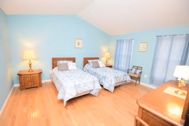 A bedroom with light blue walls and wooden flooring featuring two twin beds with matching blue and white patterned bedding. Each bed has a pillow and a small cushion. There are two bedside tables with lamps on either side of the beds. The room also contains a wooden dresser with a lamp on it, two windows with blue curtains, a wooden chair, and framed artwork on the walls.