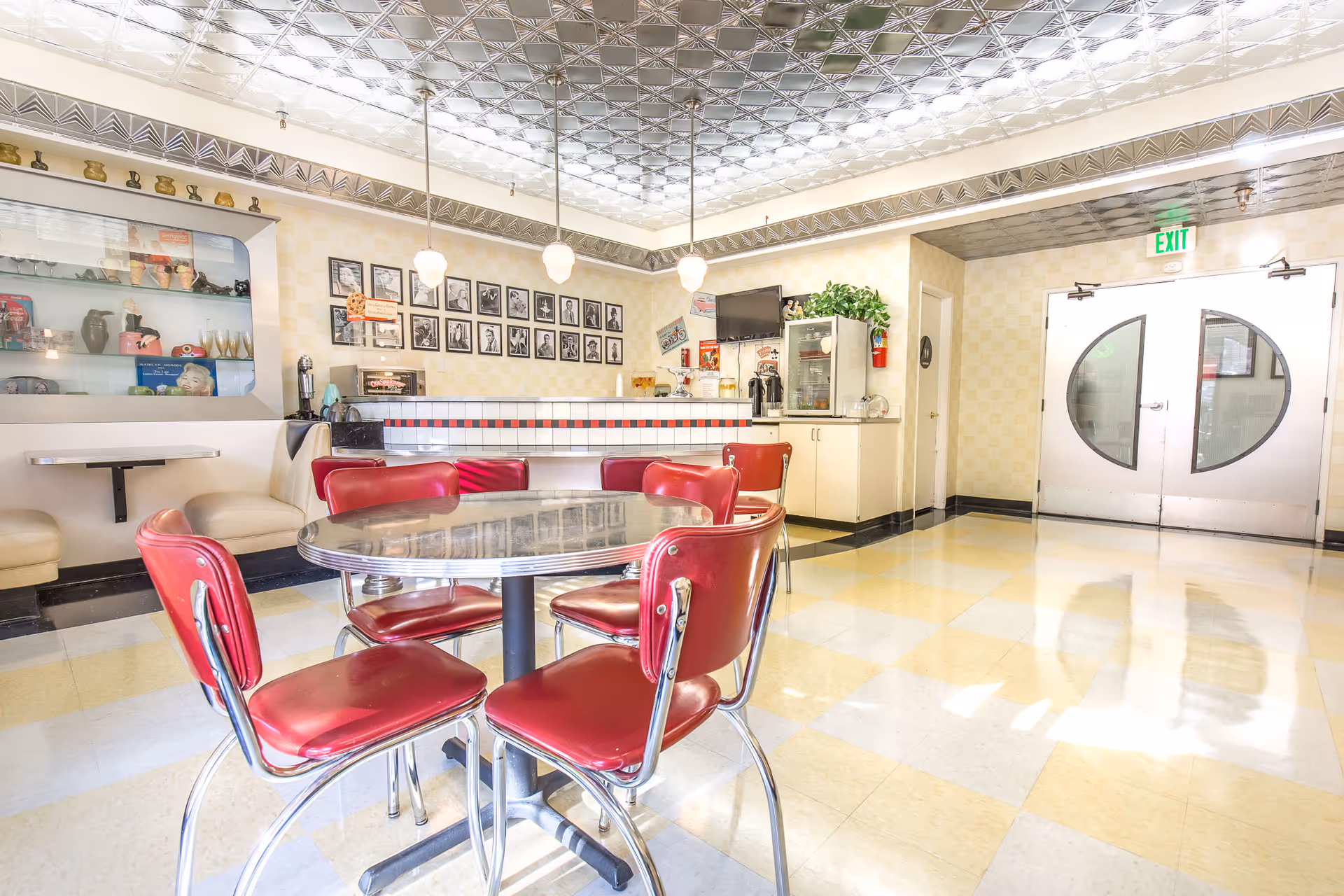Bright dining area with a round table surrounded by red cushioned chairs. The room features a retro style with a tiled floor, a counter with stools, framed black and white photos on the wall, and a display cabinet with various items. There are pendant lights hanging from a decorative ceiling and double doors with circular windows leading outside.