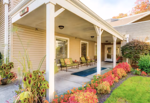 Covered porch area of a senior living facility with several green cushioned chairs along the wall, potted plants, and a colorful flower bed bordering a green lawn. The building exterior is beige with white trim and has a door at the far end of the porch.