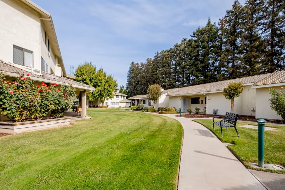 Courtyard with a curved sidewalk, benches, manicured lawn and single-story buildings set against tall trees.