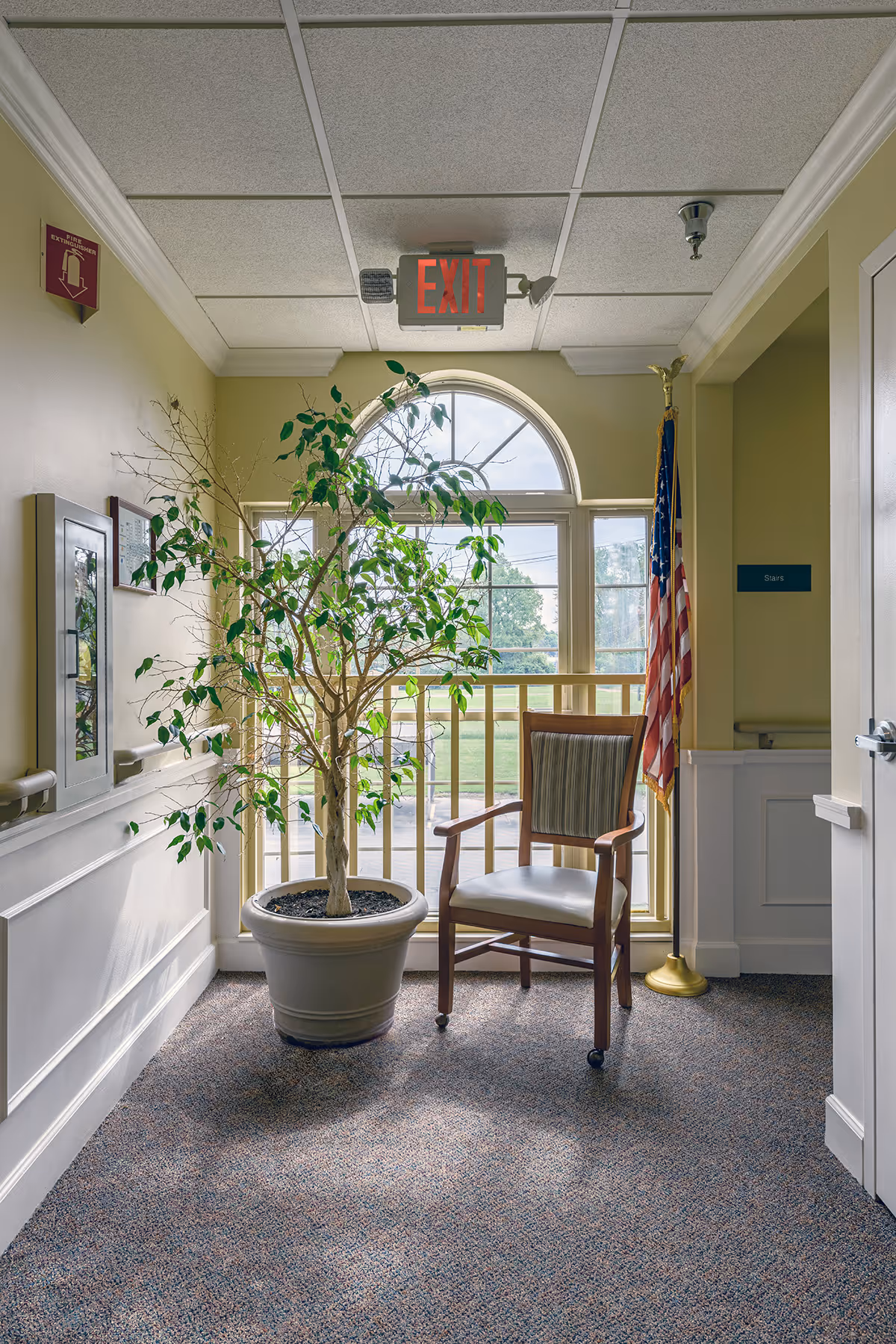 A quiet corner inside a senior living facility hallway with a large potted plant and a wooden chair with a cushioned seat and backrest on wheels. Behind them is a large arched window letting in natural light. An American flag stands to the right near a doorway labeled 'Stairs'. An illuminated exit sign hangs from the ceiling above the window.