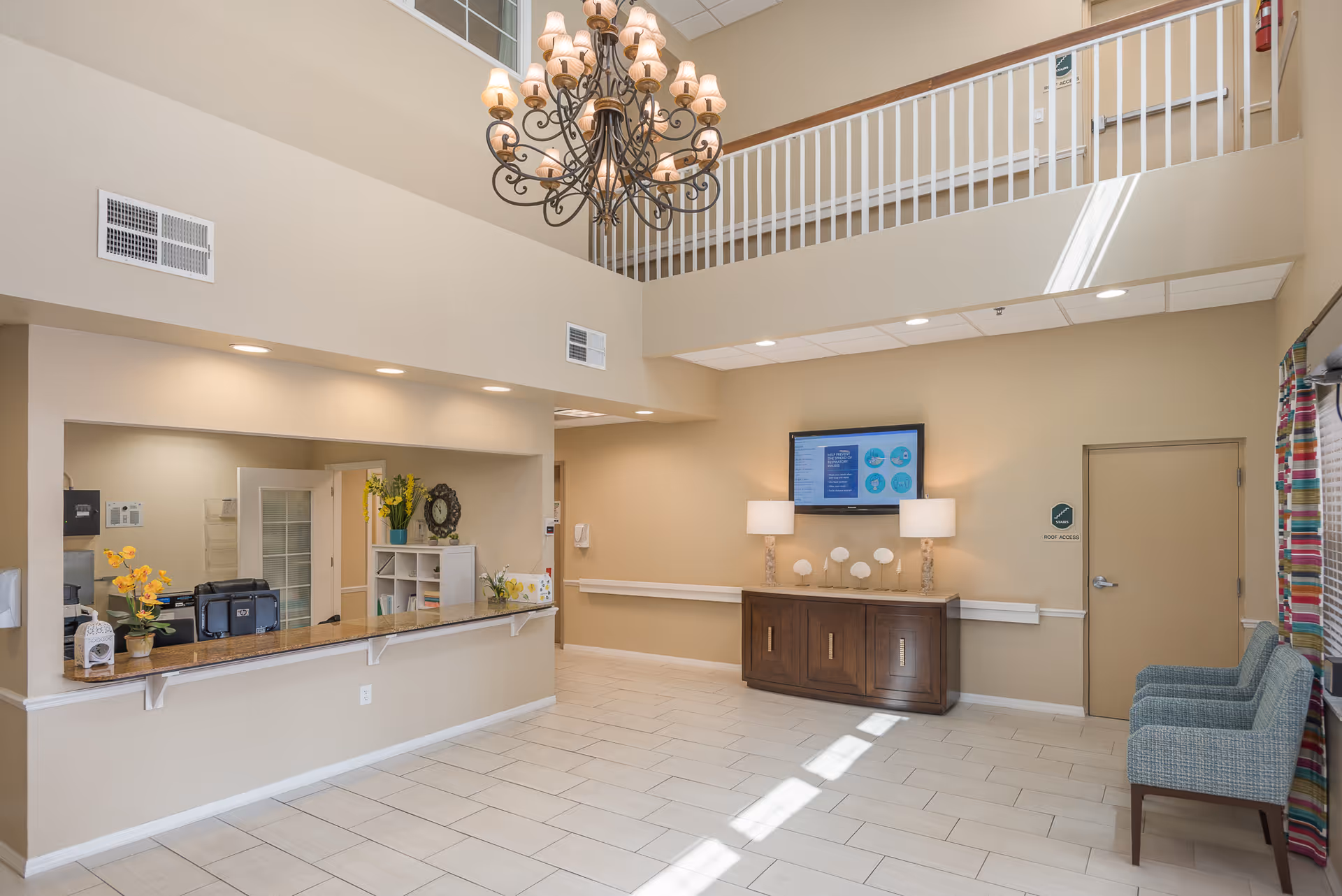 Interior view of a senior living facility lobby with a reception desk on the left, decorated with flowers and a clock. There is a large chandelier hanging from the ceiling, a wooden cabinet with two lamps and decorative items beneath a wall-mounted TV on the right. Two blue chairs are placed near a door labeled 'Roof Access'. The space has beige walls, tiled floor, and a second-floor railing visible above.