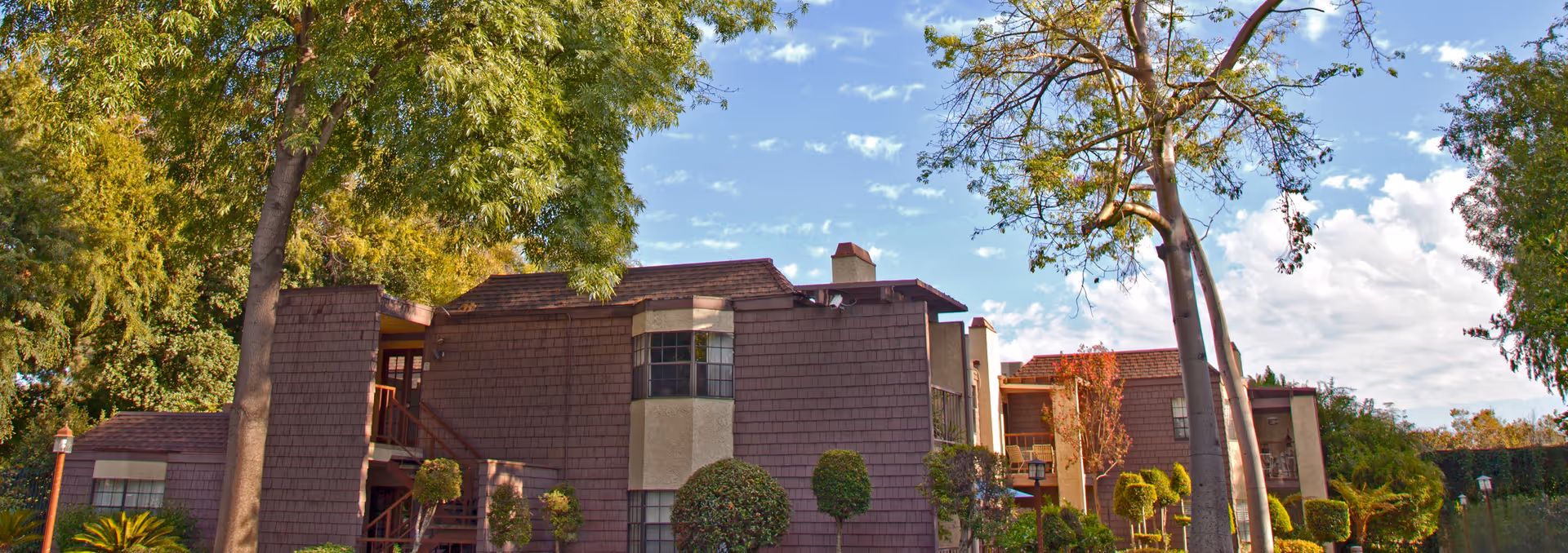 Exterior view of Oaks of Pasadena facility showing a two-story building with brown shingle siding and beige stucco accents, surrounded by well-maintained landscaping including trees, bushes, and a clear blue sky with some clouds.