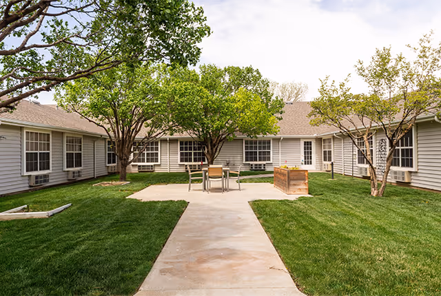 A sunny enclosed courtyard with a paved walkway, green lawn, trees, and a small table and chairs between single-story building wings.