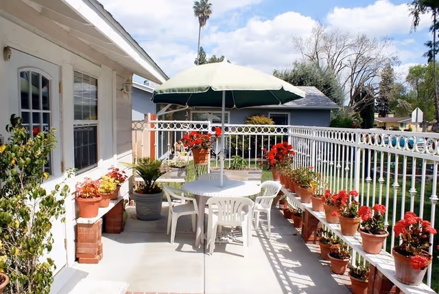 Outdoor patio area with a round white table and four white plastic chairs under a large green and white umbrella. The patio is decorated with numerous potted plants and flowers along the railing and near the house wall. The sky is partly cloudy and there are trees and neighboring houses visible in the background.