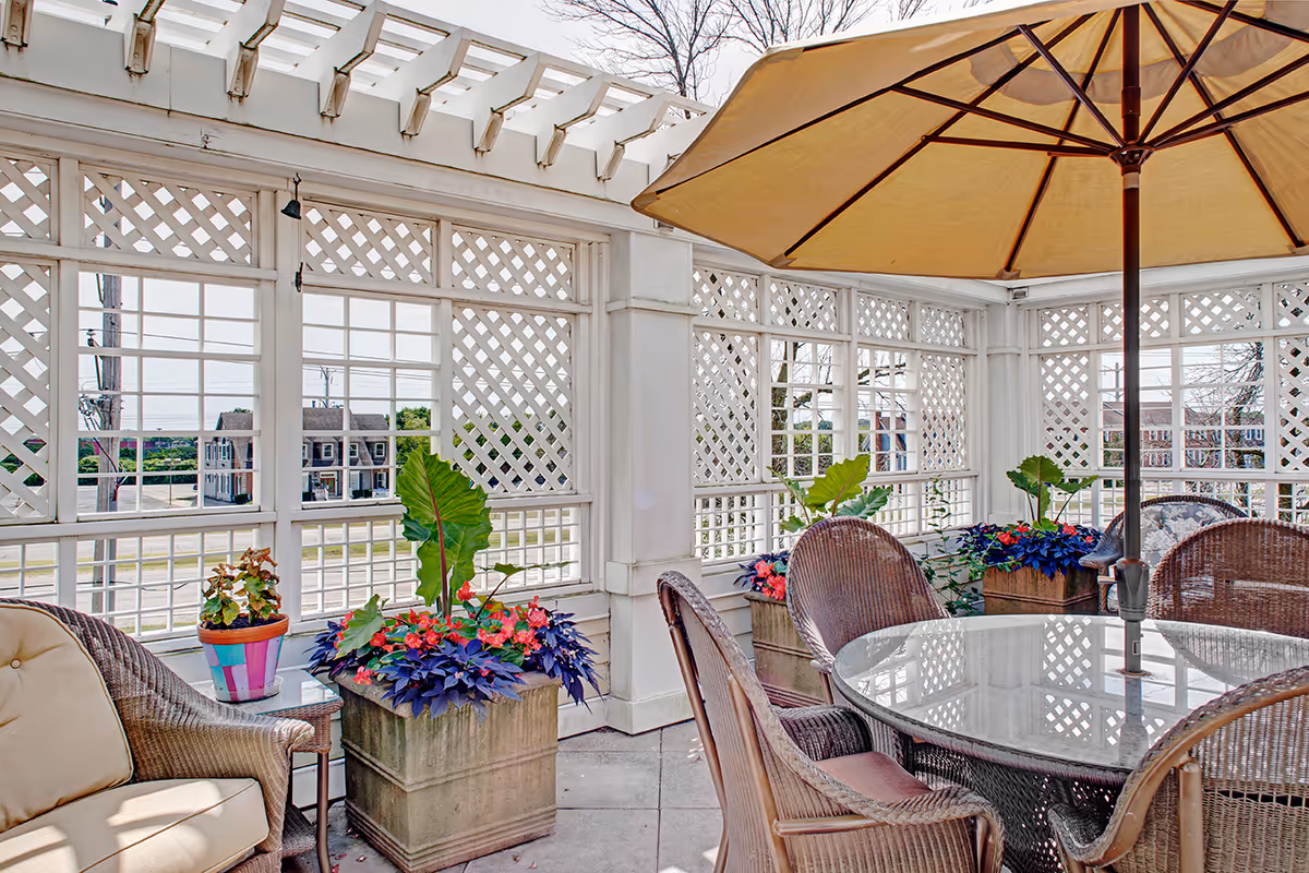 Outdoor patio area with wicker chairs and a glass-top table under a large beige umbrella. The space is enclosed with white lattice panels and decorated with potted plants and flowers.