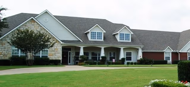 Exterior view of a single-story senior living facility building with a combination of stone and siding facade, a covered front porch with columns, multiple windows including dormer windows on the roof, and a well-maintained lawn in front.