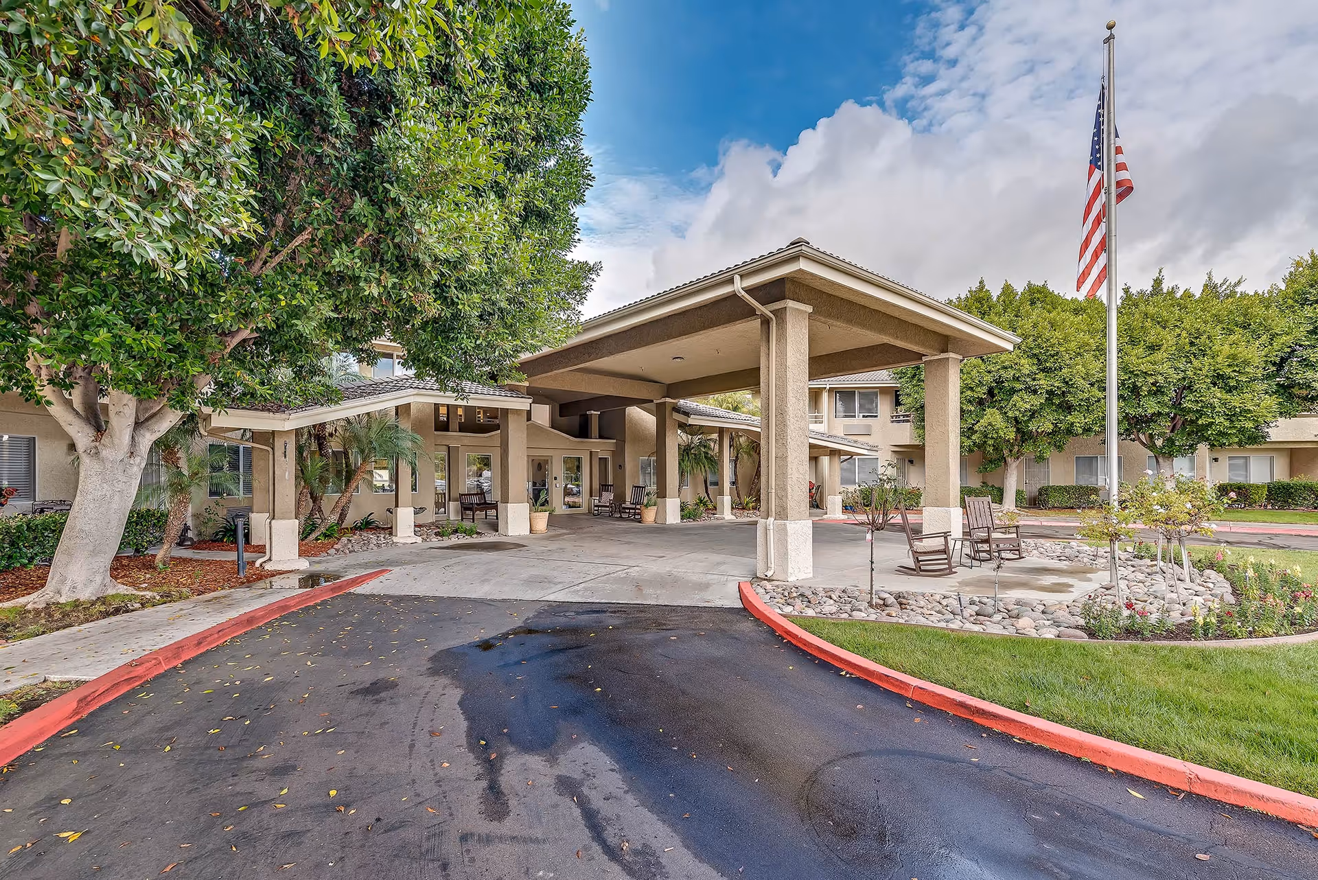 Front exterior view of Arcadia Place Senior Living facility showing a covered entrance with columns, a driveway, trees, an American flag on a flagpole, and outdoor seating with chairs and a table.