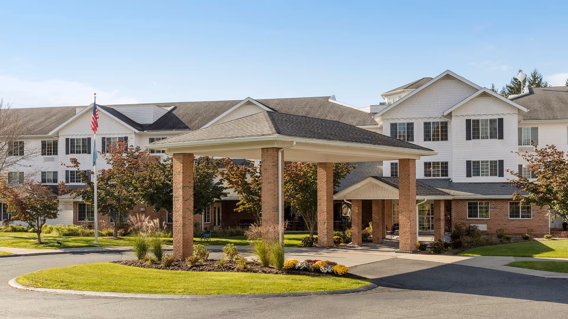 Front exterior view of Holiday Bluebird Estates, a multi-story senior living facility with a covered entrance supported by brick columns, surrounded by landscaped greenery and trees under a clear blue sky.