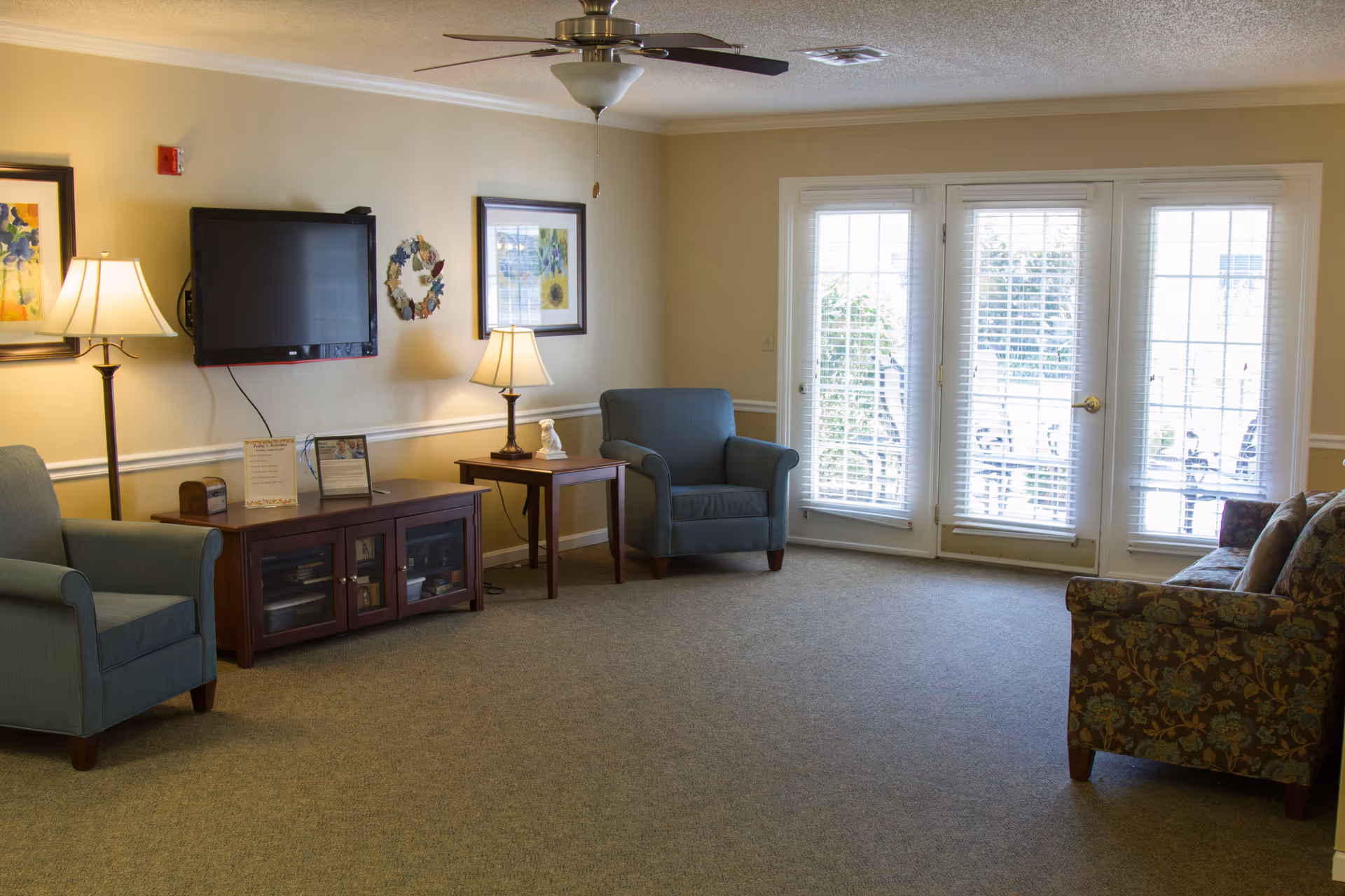 A cozy living room with beige walls and carpeted floor. The room features a ceiling fan with light, two blue armchairs, a floral-patterned sofa, a wooden TV stand with a flat-screen TV mounted above it, two table lamps, framed floral artwork on the walls, and large glass doors with blinds leading outside.