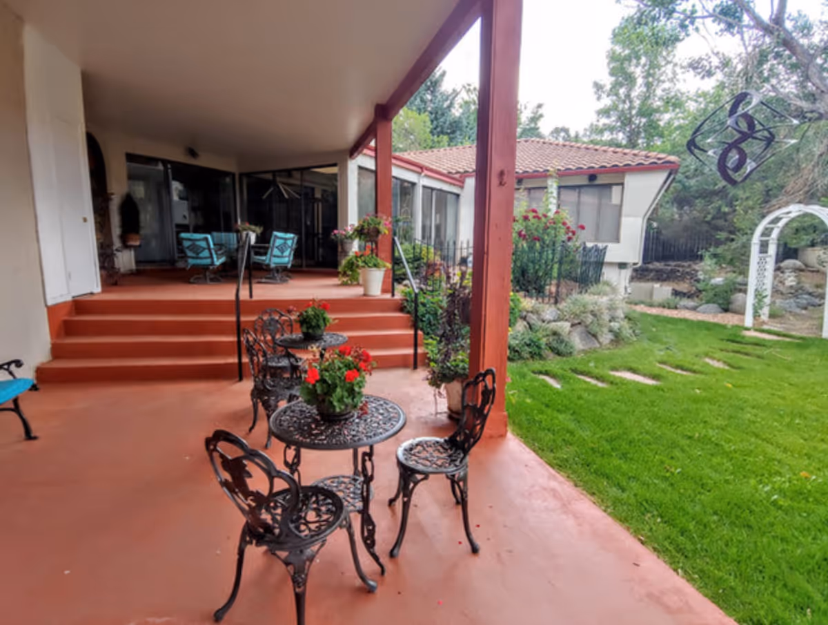 Covered patio with wrought-iron tables and chairs, potted flowers, steps up to a seated area and a green lawn with a garden arch.