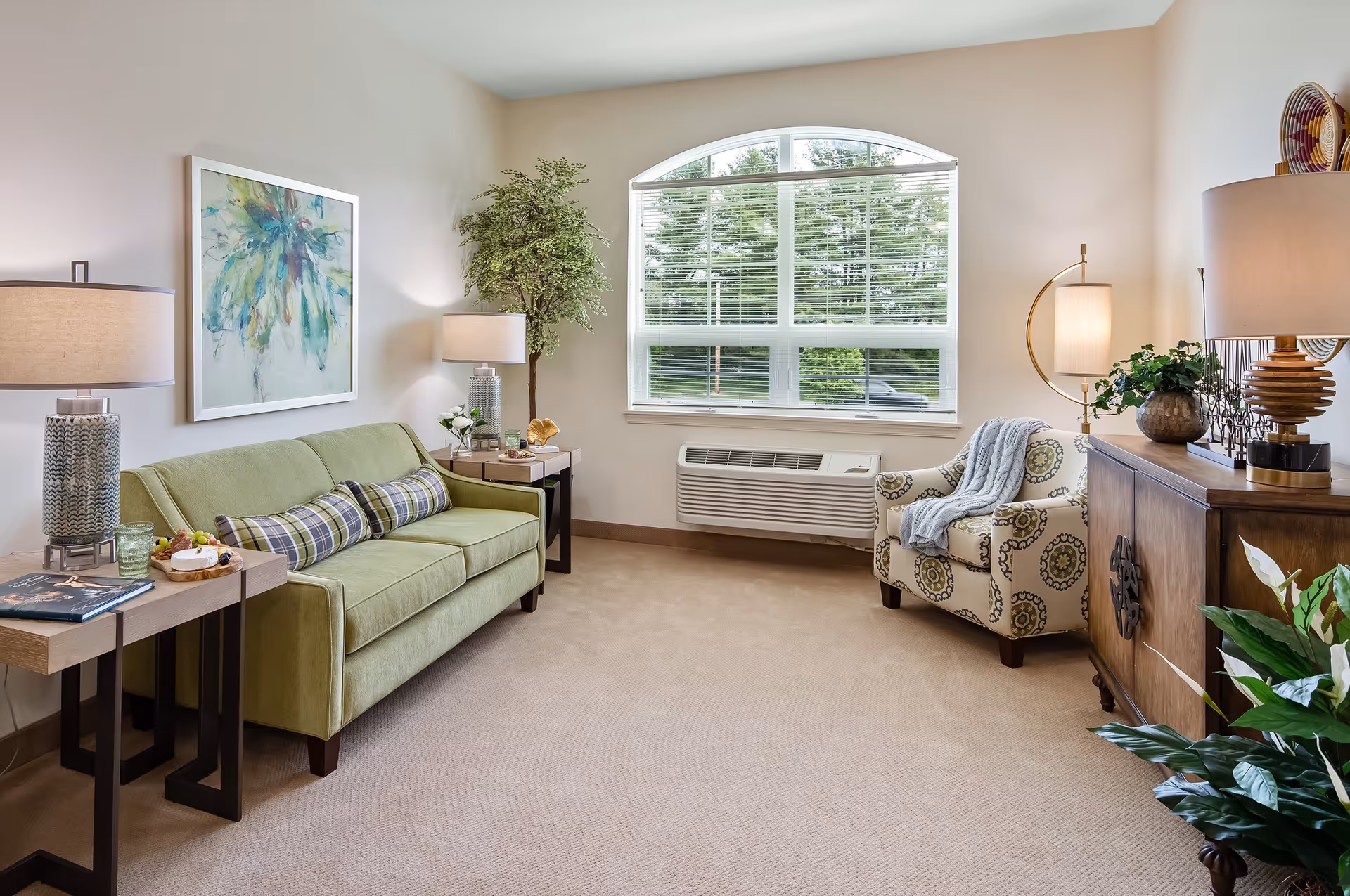 Bright furnished living room with a green sofa, patterned armchair, lamps, large window, and decorative plants.