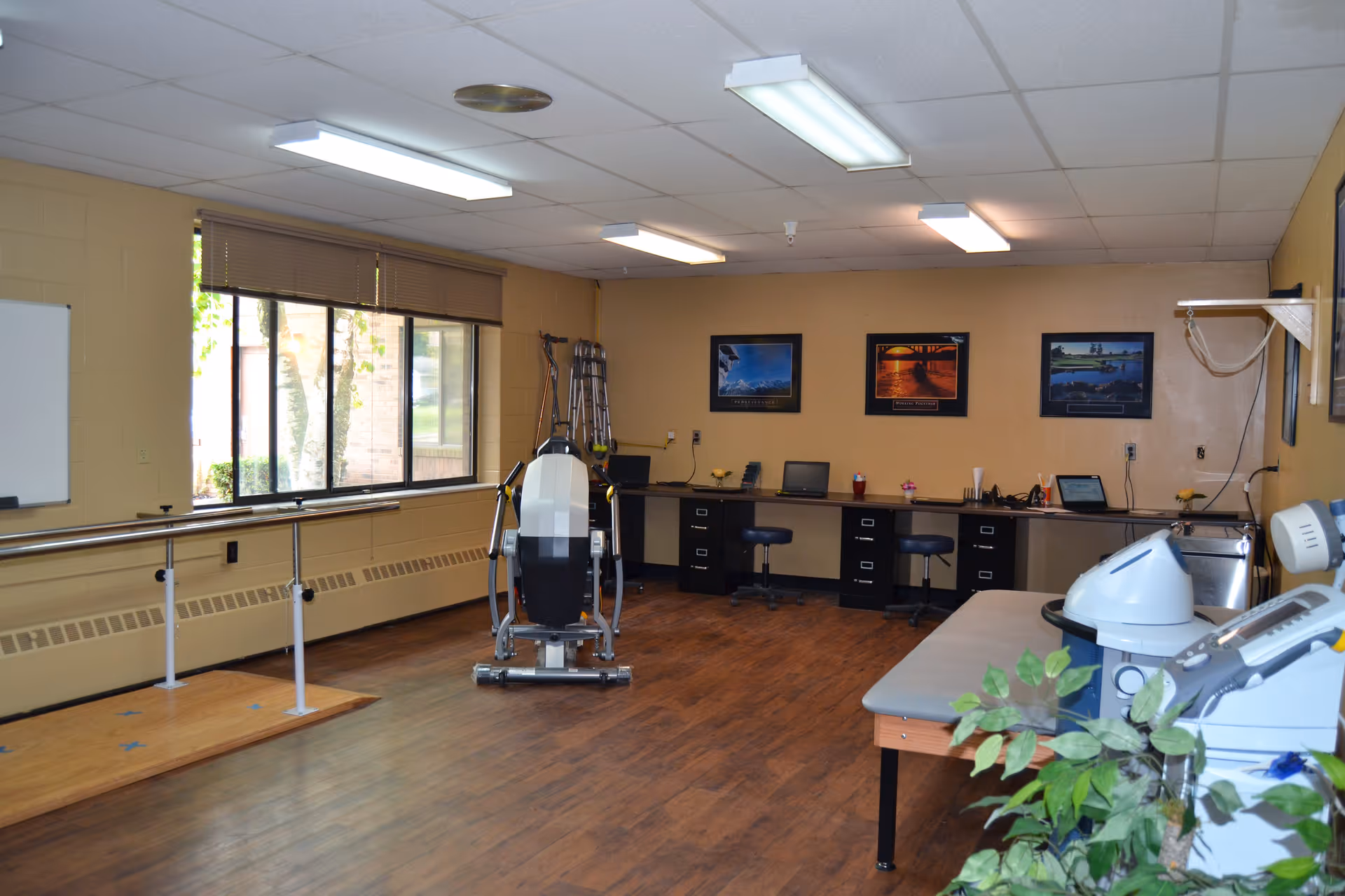 A spacious rehabilitation therapy room with parallel bars, exercise equipment, desks and a treatment table.