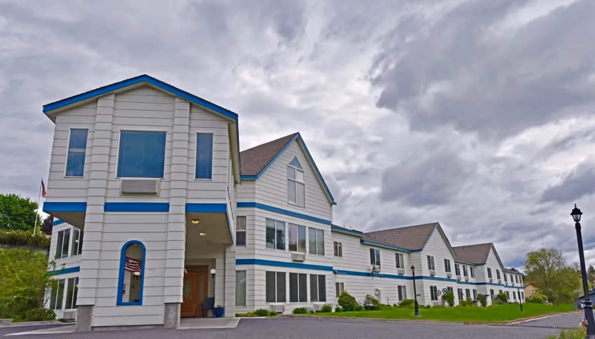 Exterior view of a large two-story senior living facility building with white siding and blue trim under a cloudy sky. The building has multiple windows, a covered entrance, and a well-maintained lawn with street lamps along the driveway.