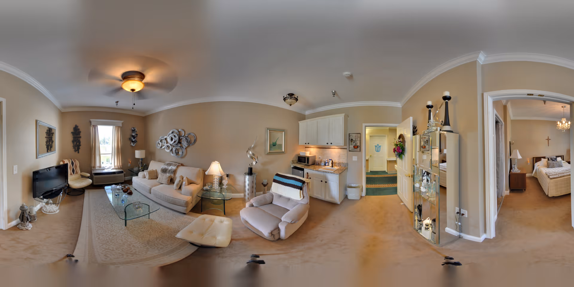 A spacious and well-lit senior living apartment featuring a living room with a beige sofa, armchair, glass coffee table, and a TV on a stand. The room has beige walls and carpet, with decorative wall art and a window with curtains. Adjacent to the living room is a small kitchenette with white cabinets, a microwave, and a countertop. To the right, an open doorway leads to a bedroom furnished with a bed, nightstands, lamps, and a cross on the wall. The overall decor is warm and inviting with neutral tones.