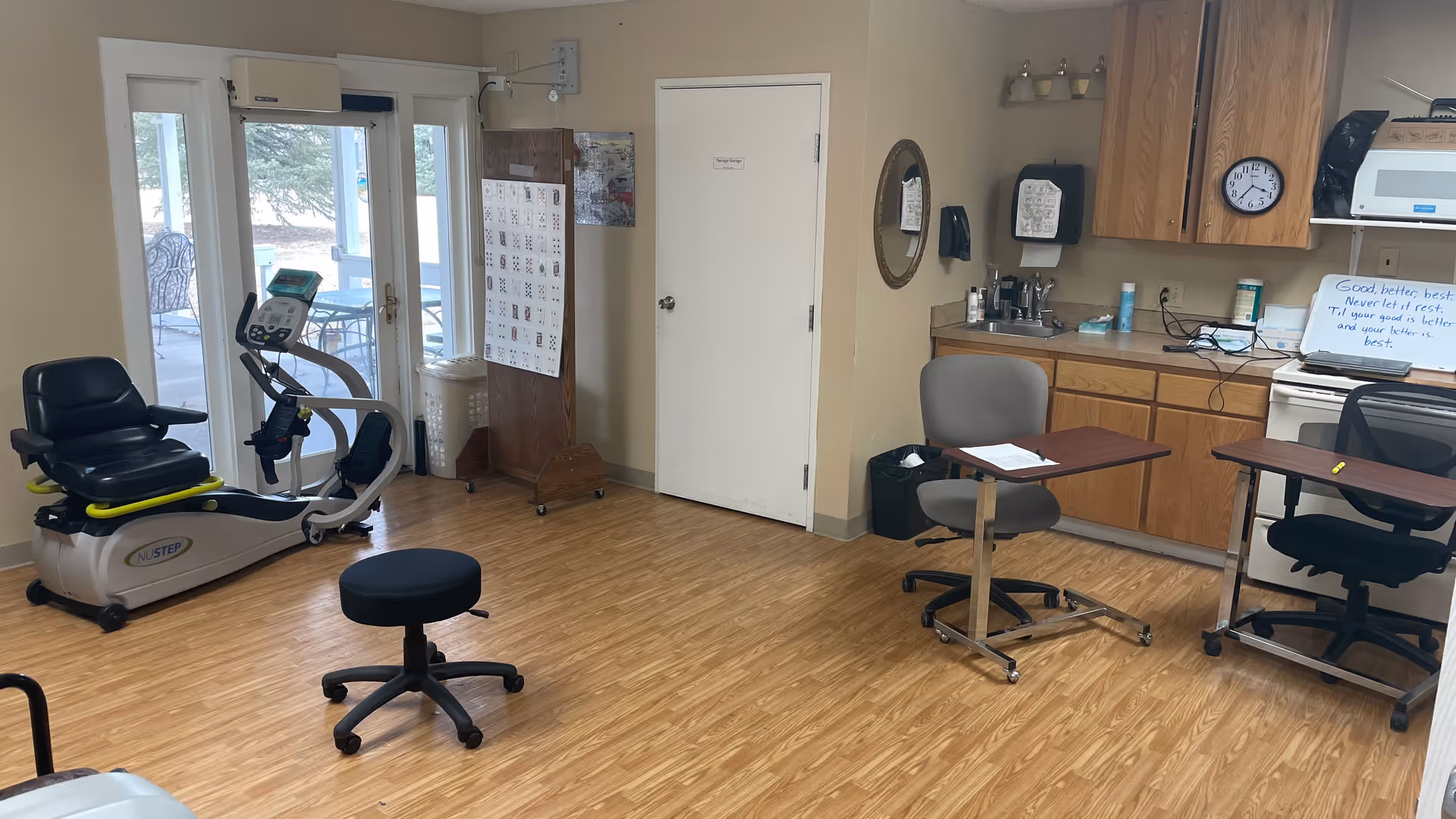 A therapy room with exercise equipment including a NuStep recumbent cross trainer, two chairs with attached rolling desks, a small rolling stool, and a kitchenette area with wooden cabinets, a sink, microwave, and a clock on the wall. There is a door labeled 'Therapy Storage' and a whiteboard with a motivational quote. Glass doors lead to an outdoor patio area.