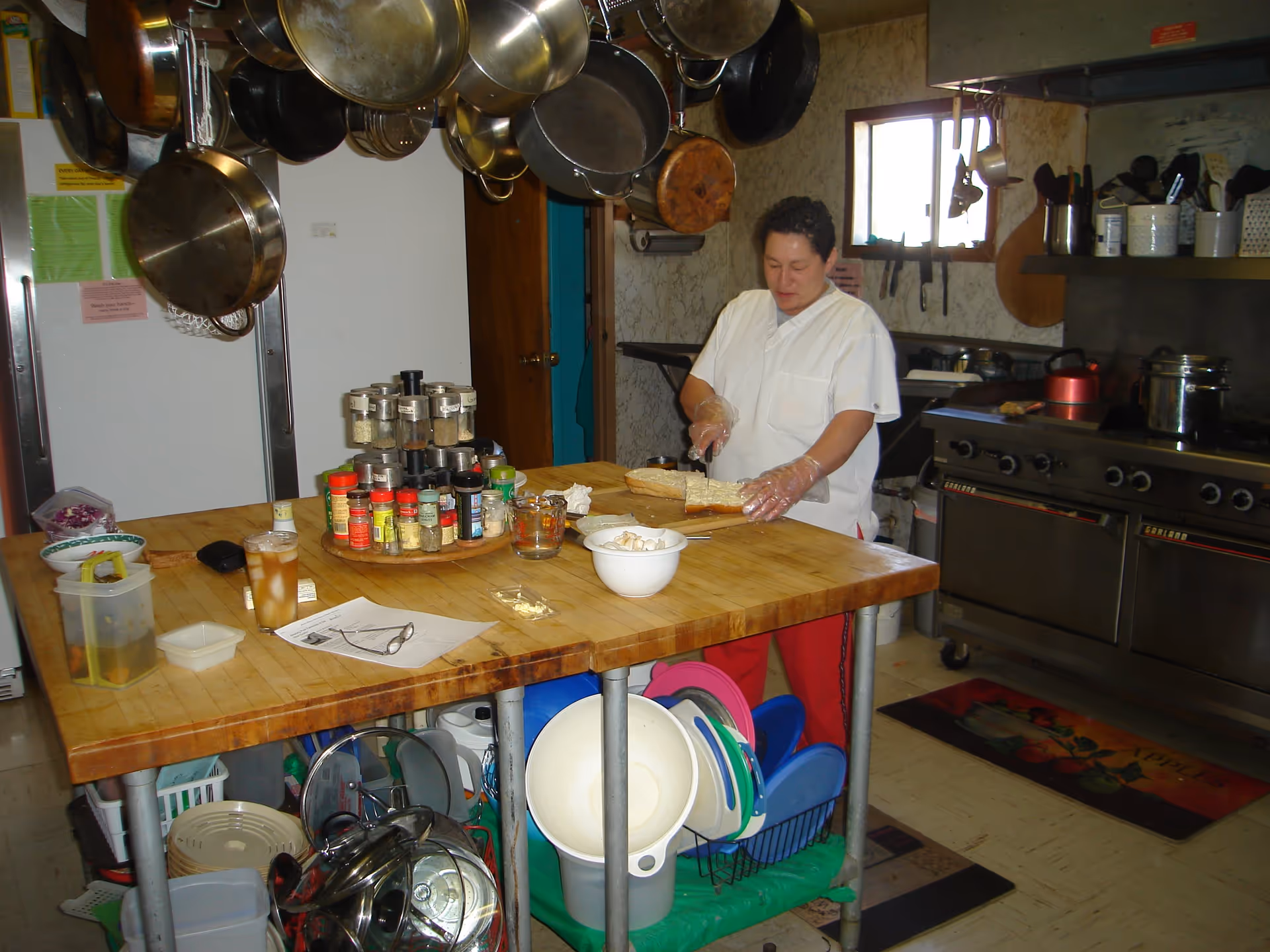 A person wearing a white chef coat and red pants is slicing a loaf of bread on a wooden kitchen island. The kitchen has hanging pots and pans above the island, various spices on the counter, and a large stove with pots and utensils. There are kitchen tools and dishes stored under the island, and a window on the wall behind the person.