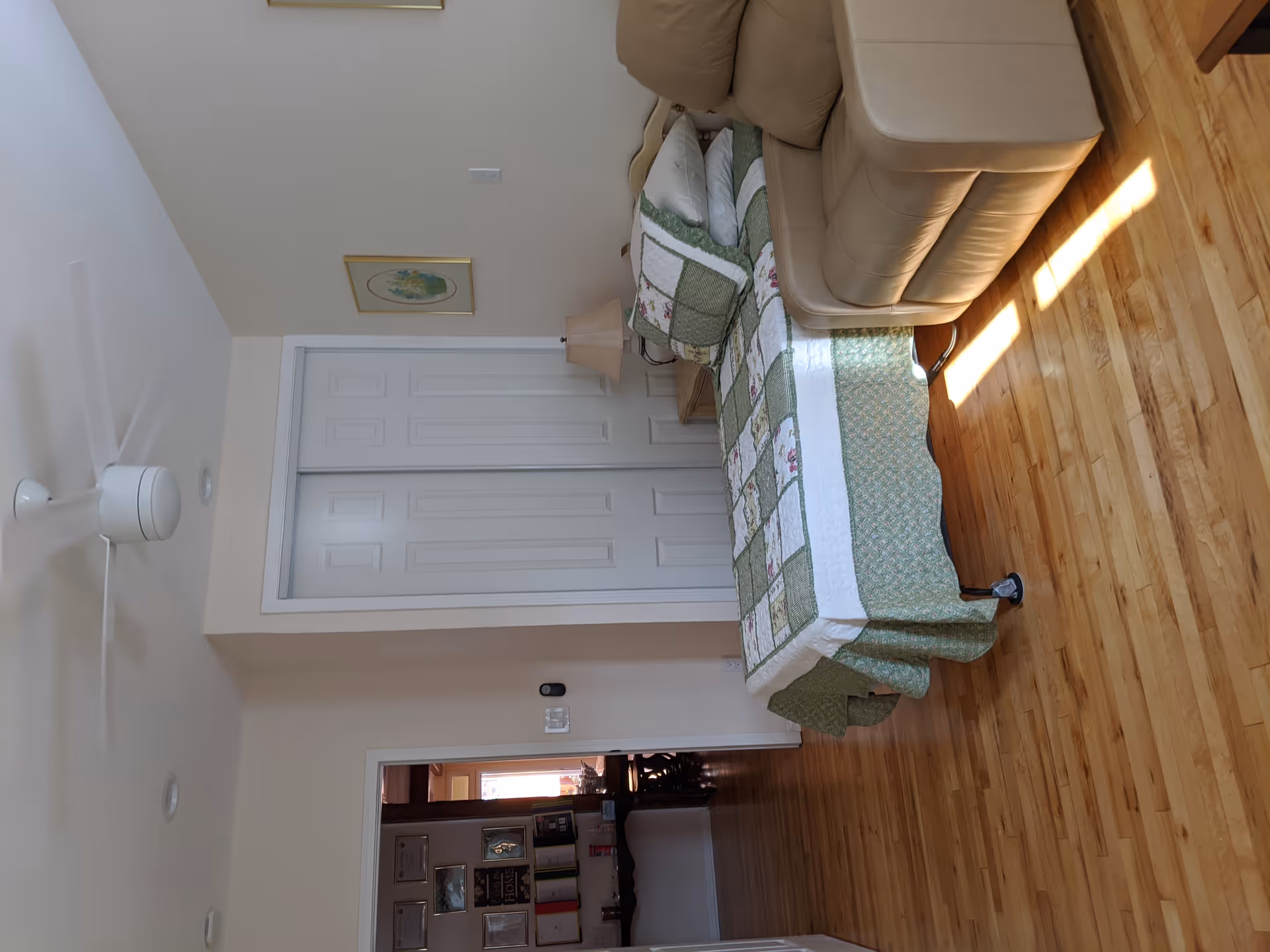 A bedroom with a single bed covered in a green and white patchwork quilt, a beige recliner chair next to the bed, a wooden nightstand with a lamp, a white ceiling fan, and a closet with white sliding doors. The room has light-colored walls and wooden flooring.