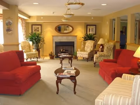 A cozy living room with two red sofas, two floral armchairs, a striped armchair, and two wooden coffee tables. The room features a fireplace with a mirror above it, two framed pictures on the wall, potted plants, and warm lighting from ceiling fixtures and a table lamp.