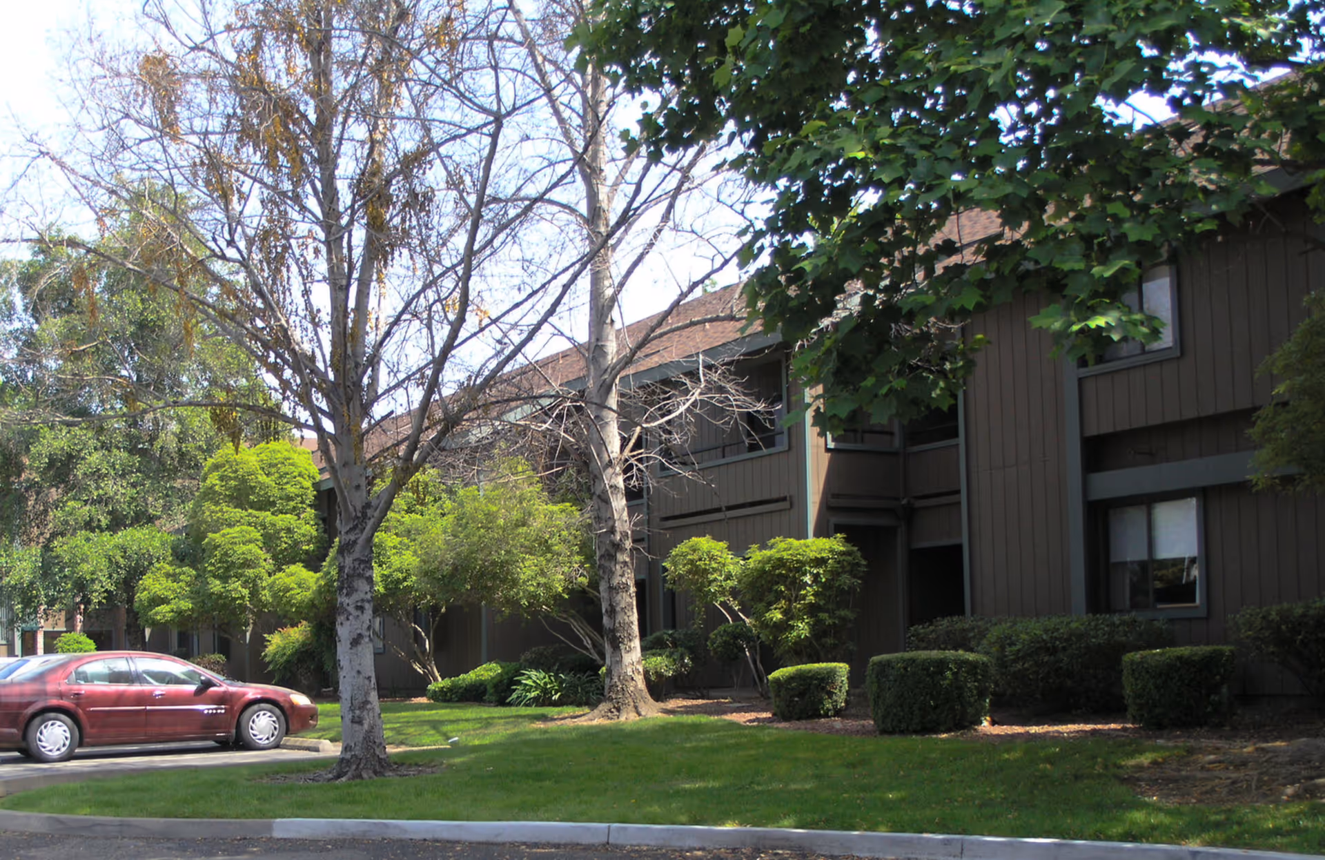 Two-story brown residential building with landscaped shrubs and trees and a red car parked in front.