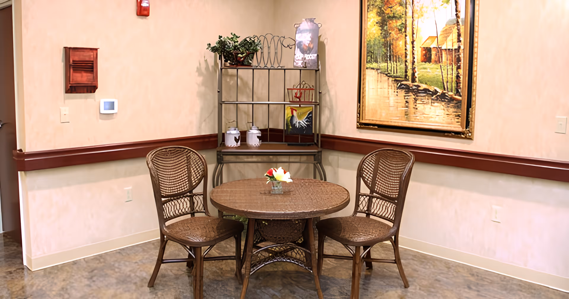A small seating area with a round wicker table and three matching chairs against a decorated wall with shelving and a framed painting.