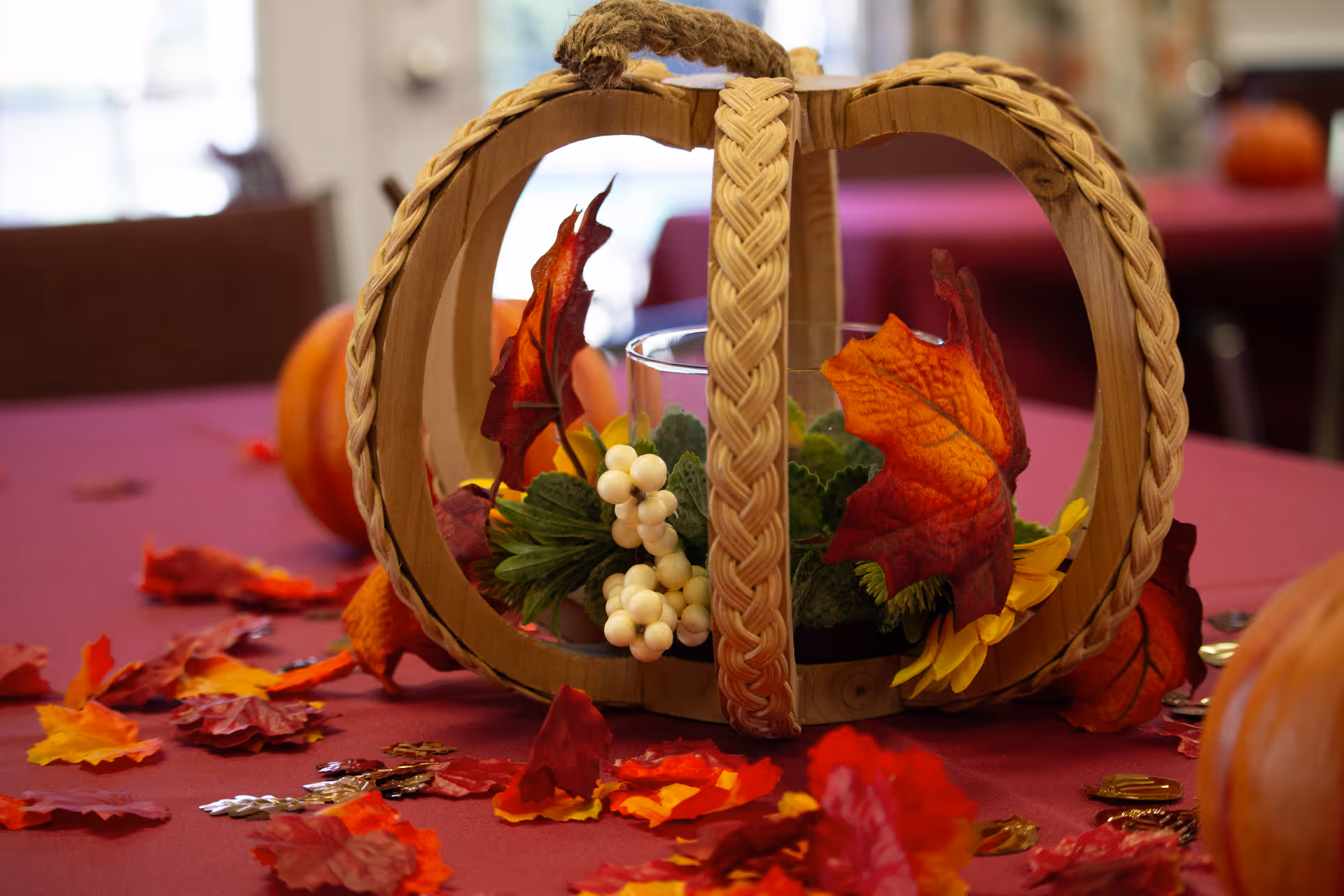 Autumn-themed table centerpiece featuring a woven wooden pumpkin filled with faux leaves, berries and a candle on a burgundy tablecloth.