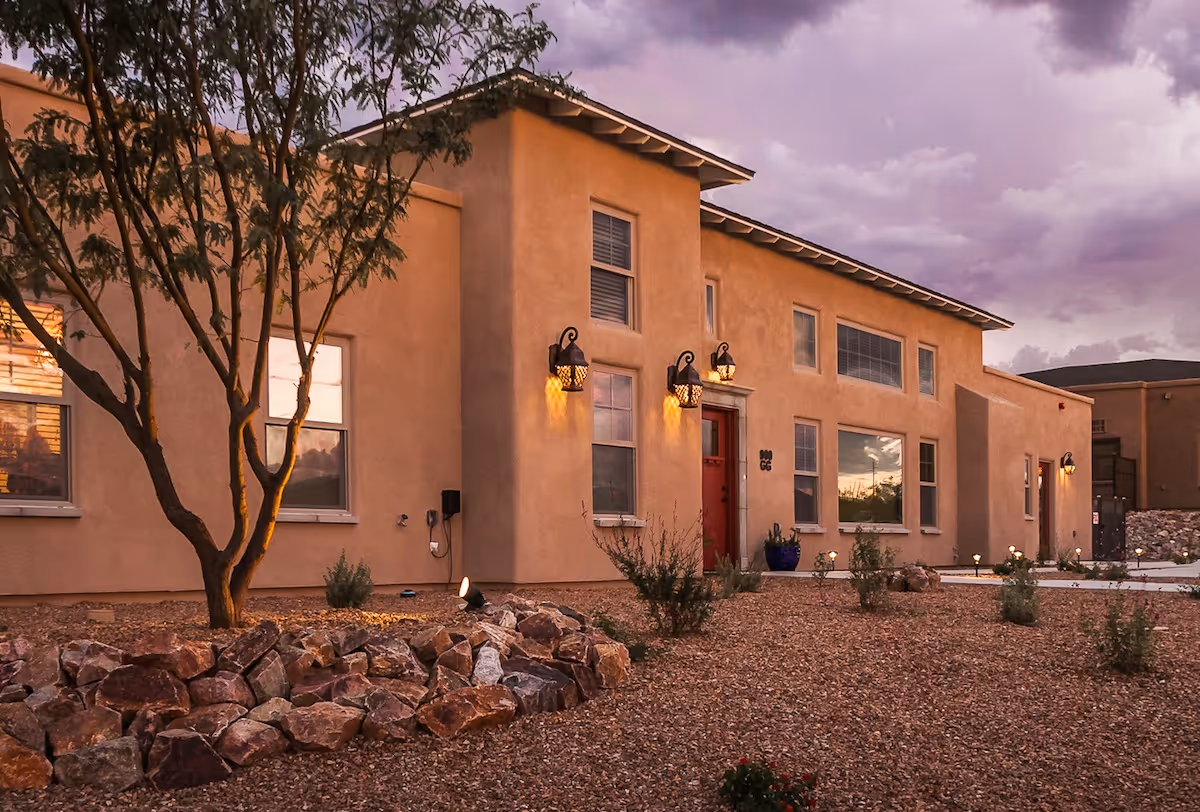 Front exterior of a stucco building with desert rock landscaping, illuminated lanterns, and a red entrance door at dusk.
