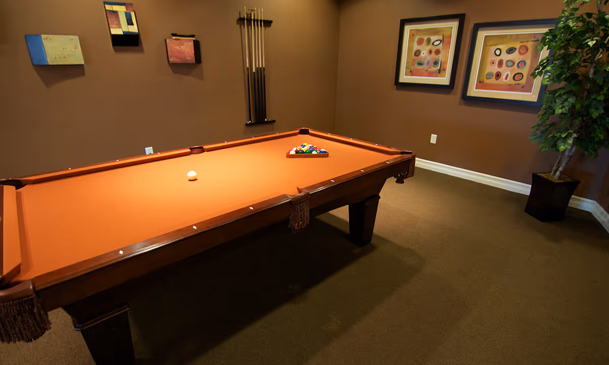 A billiards table with orange felt in a brown-walled game room with pool cues, framed artwork, and a potted plant.