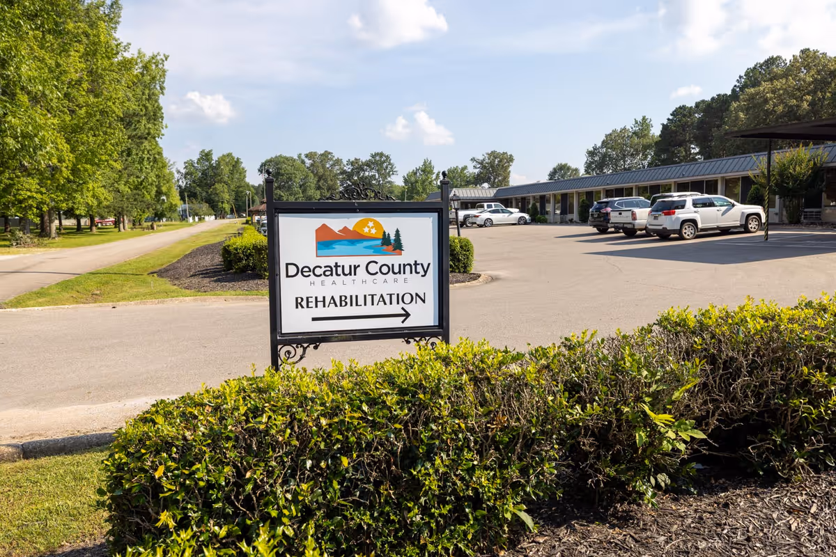 Sign reading 'Decatur County Healthcare Rehabilitation' in front of a parking lot and single-story facility under a sunny sky.