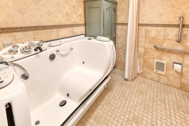 A bathroom featuring a large white walk-in bathtub with multiple control knobs and a handheld showerhead. The walls are tiled halfway up with beige tiles and have a patterned wallpaper above. There is a green cabinet in the corner and a beige curtain hanging beside the tub. The floor is covered with small hexagonal beige tiles. Safety grab bars and a toilet paper holder are mounted on the wall.