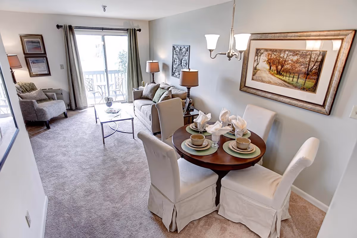 A cozy living and dining area in a retirement facility with a round wooden dining table set for four with plates, cups, and napkins. The living area has a beige sofa, two lamps, a coffee table, and an armchair near a sliding glass door leading to a balcony with outdoor chairs. The walls are decorated with framed artwork and a metal wall hanging.