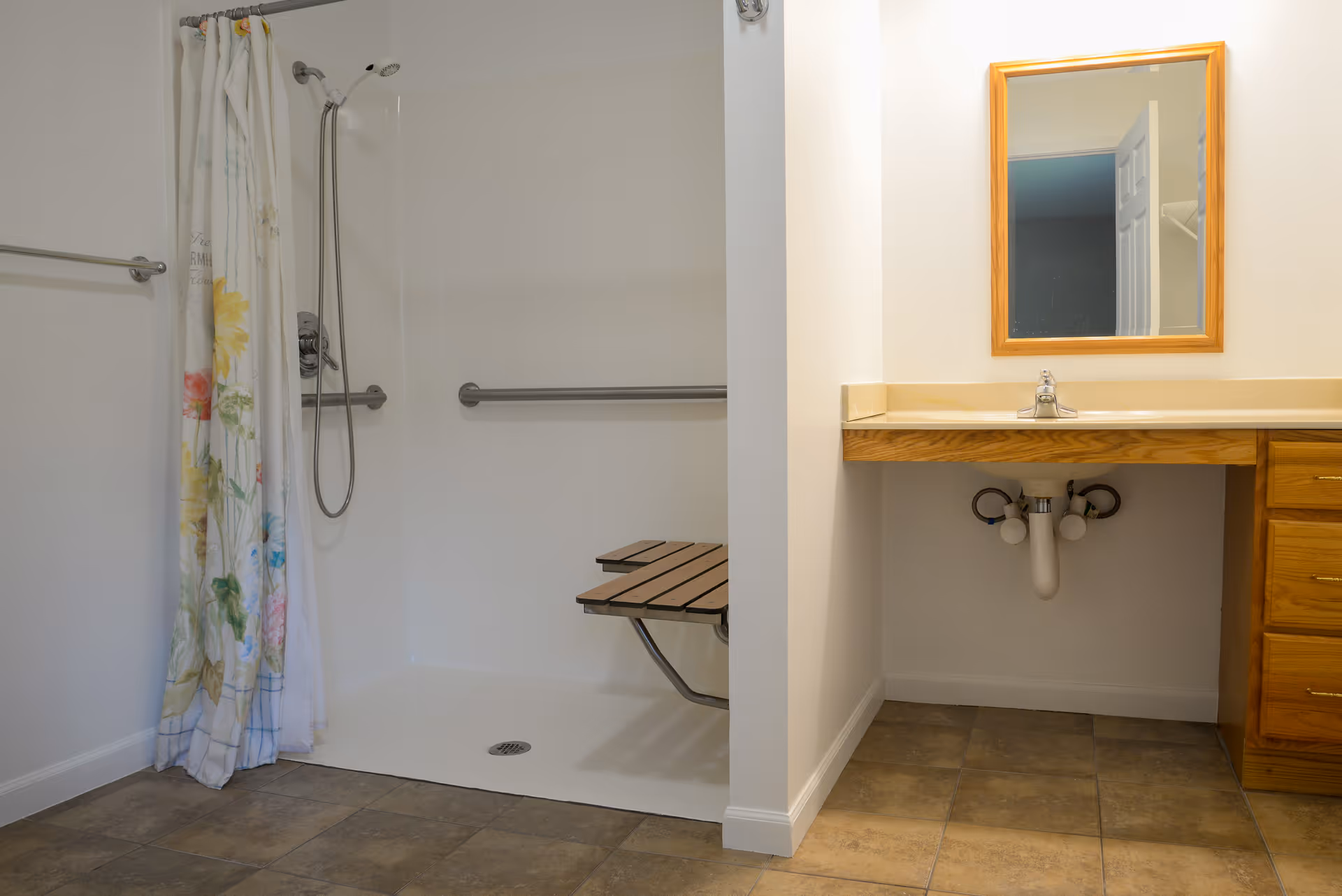 A bathroom with a walk-in shower featuring a floral shower curtain and a foldable wooden shower seat. There are grab bars installed on the shower walls. Next to the shower is a vanity with a sink, a wooden framed mirror, and wooden drawers. The floor is tiled.