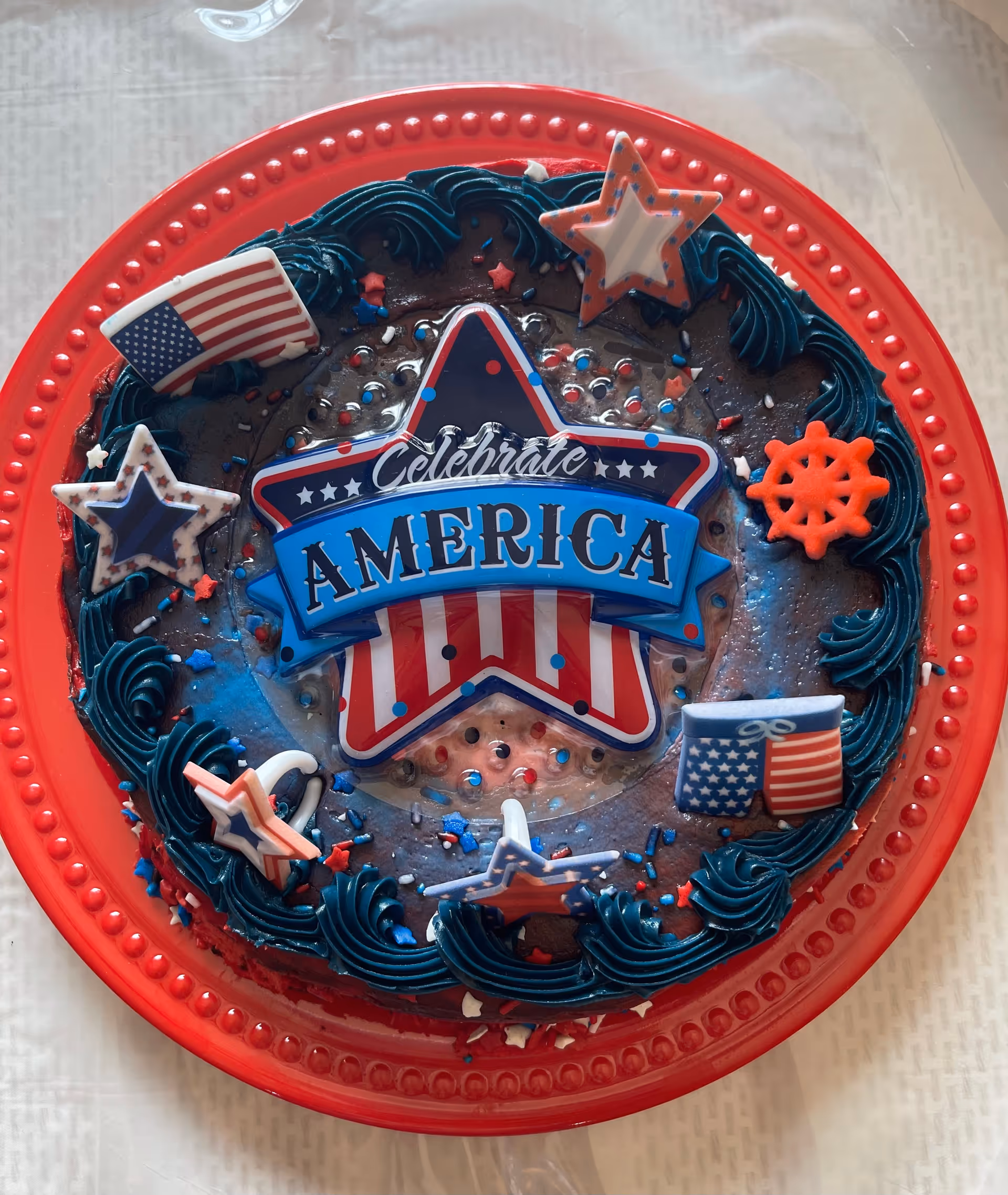 A round chocolate cake decorated with blue frosting and patriotic-themed decorations including stars, an American flag, and a sign that says 'Celebrate America' on a red plastic tray.
