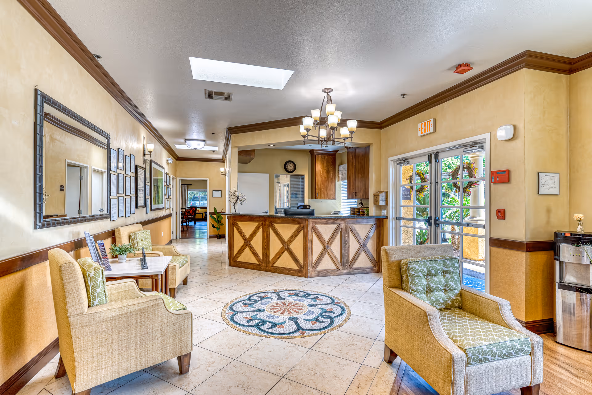 Bright lobby-style reception area with a wooden front desk, upholstered chairs, chandelier, and tiled floor with decorative medallion.