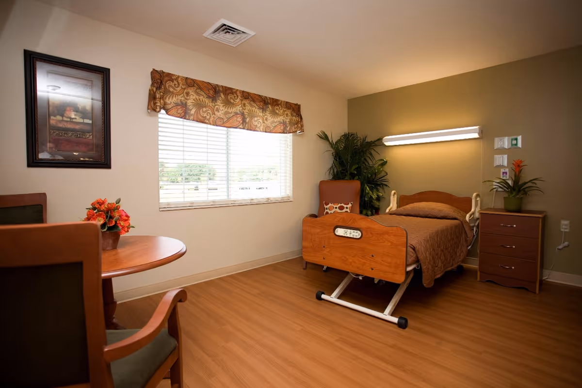 A furnished senior care bedroom with an adjustable wooden hospital bed, nightstand, chair, round table and window with blinds.