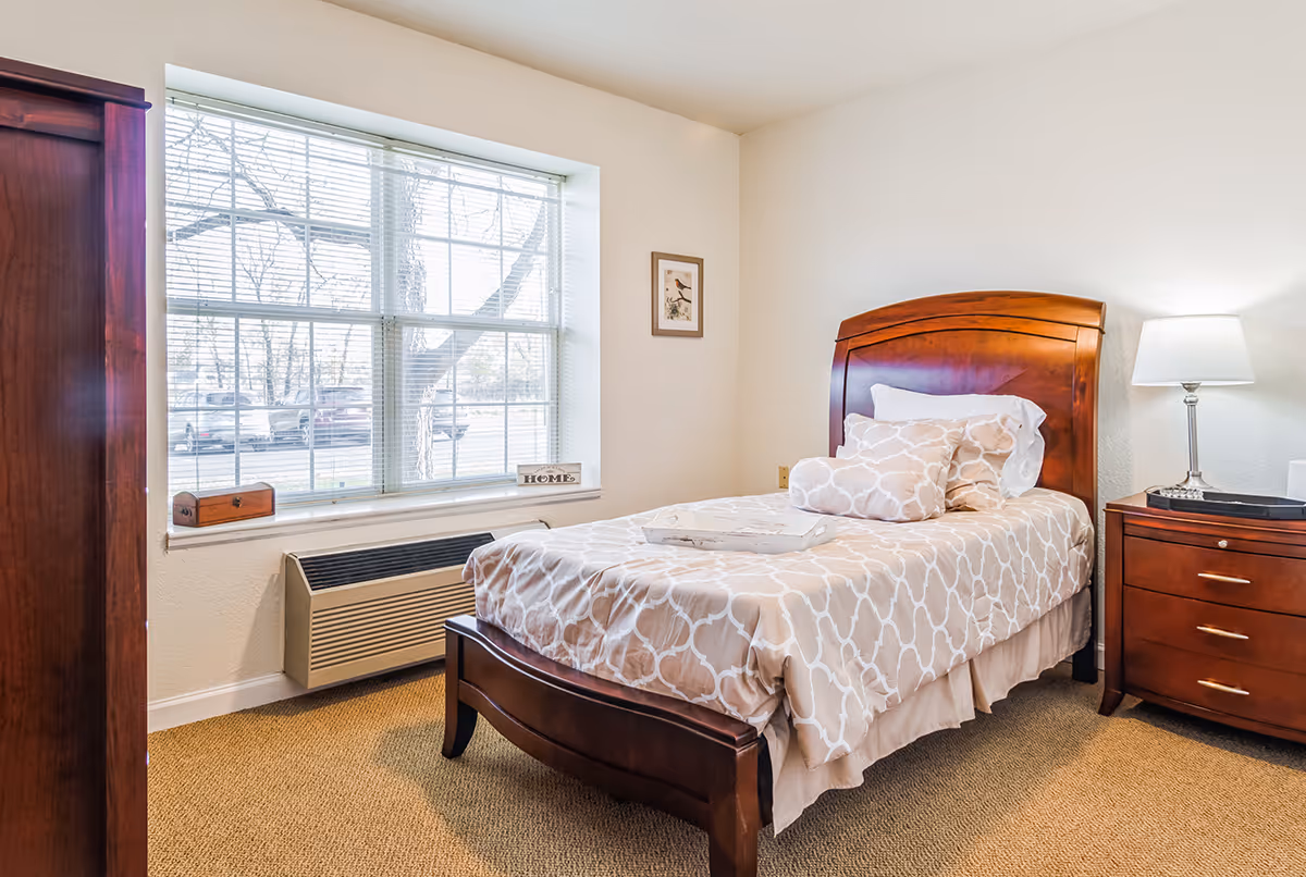 A cozy bedroom with a single wooden bed featuring beige patterned bedding and multiple pillows. Next to the bed is a wooden nightstand with a lamp and a tray on the bed. A large window with blinds lets in natural light, and a small framed picture hangs on the wall. The room has beige carpet and light-colored walls.