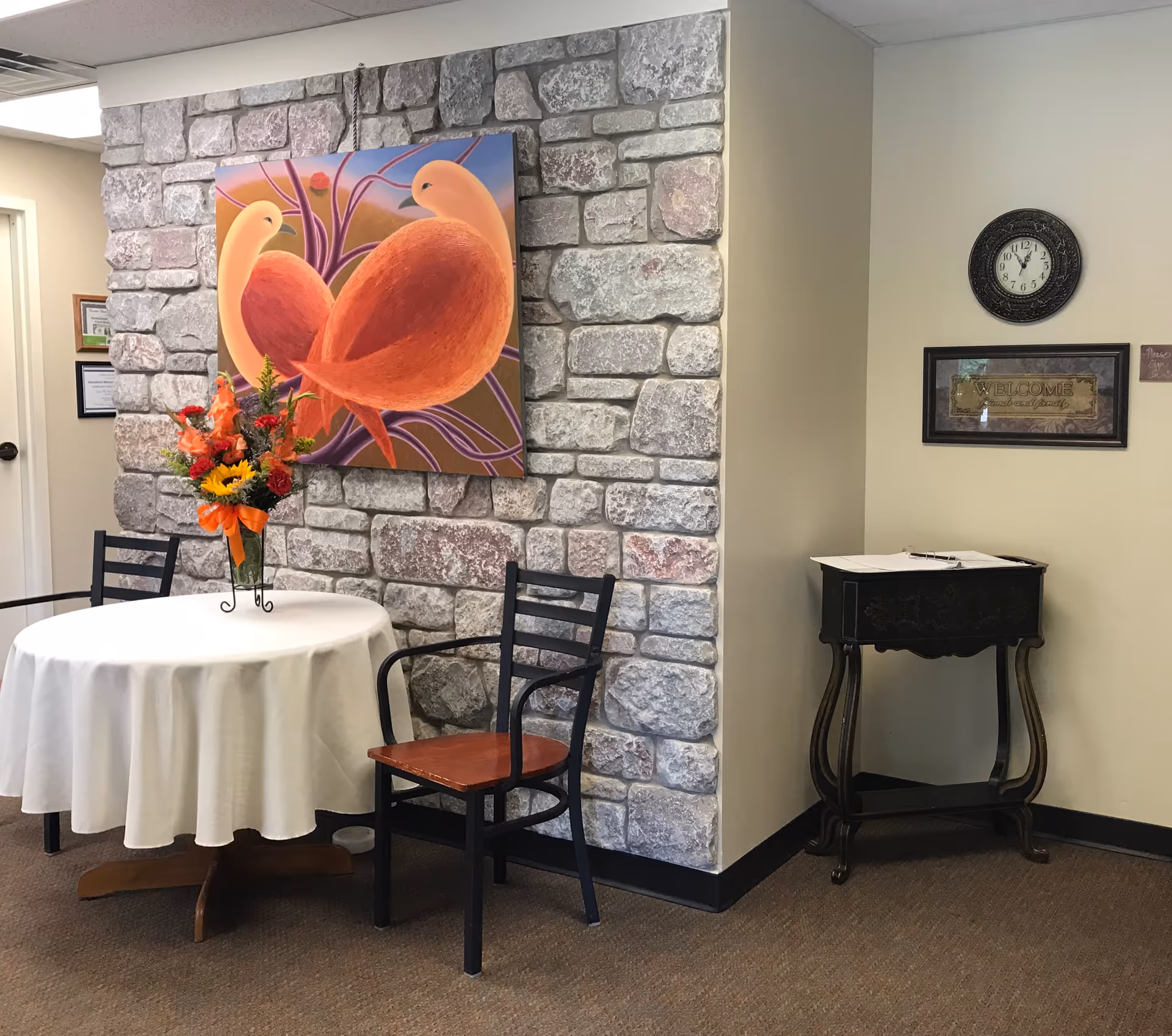 A small seating area with a round table covered with a white tablecloth and two black chairs with wooden seats. On the table is a vase with a colorful flower arrangement. Behind the table is a wall with a stone facade and a large painting of two abstract orange birds. To the right, there is a small black wooden stand with a guestbook on top, a framed 'Welcome' sign, and a round black clock on the wall showing 12:05.