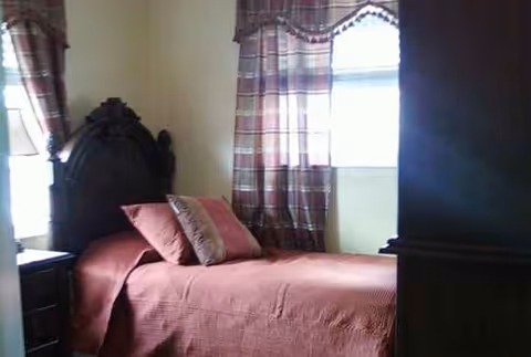 A furnished bedroom with a dark wood headboard, pink bedding and plaid curtains.