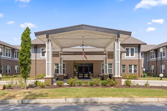 Front entrance of a two-story brick senior living building with a covered porte-cochere, American flag, and landscaped lawn.