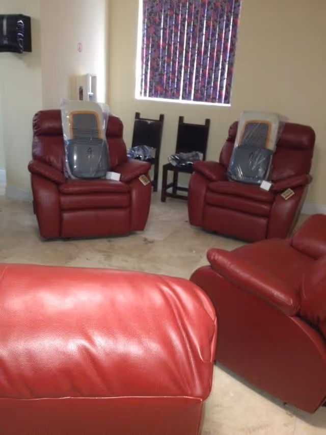 A room with four red leather recliner chairs arranged in a semi-circle. Two of the chairs have plastic coverings on the seat and backrest. Behind the chairs, there are two dark wooden chairs and a small table. A window with vertical blinds featuring a colorful pattern is visible on the wall.