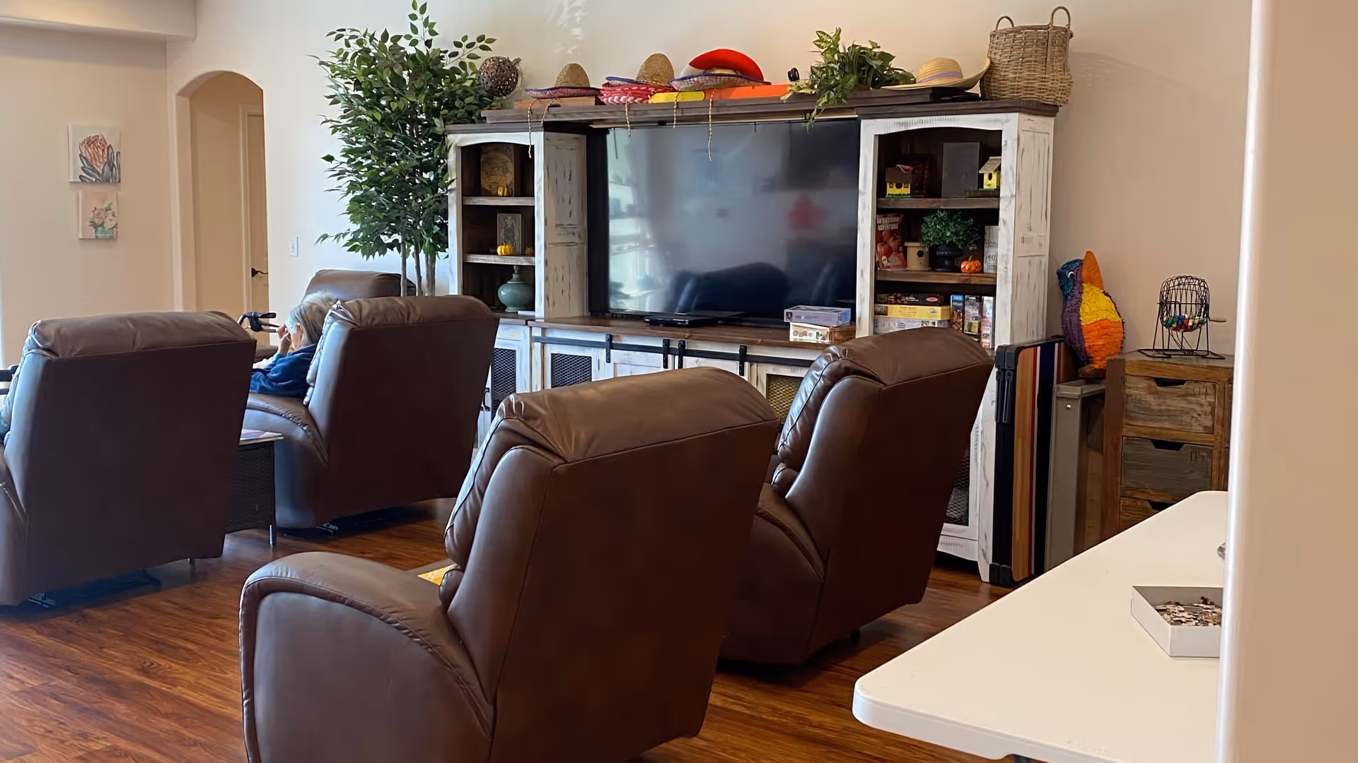 Living room with several brown leather recliners arranged facing a large flat-screen TV in a white entertainment center with plants and shelving.