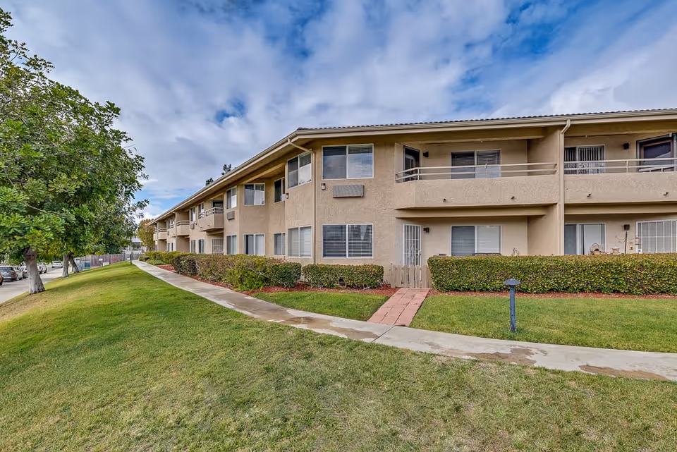 Exterior view of a two-story beige senior living facility building with multiple windows and balconies. There is a well-maintained green lawn with a concrete walkway leading to the entrance. Trees and bushes line the side of the building under a partly cloudy sky.