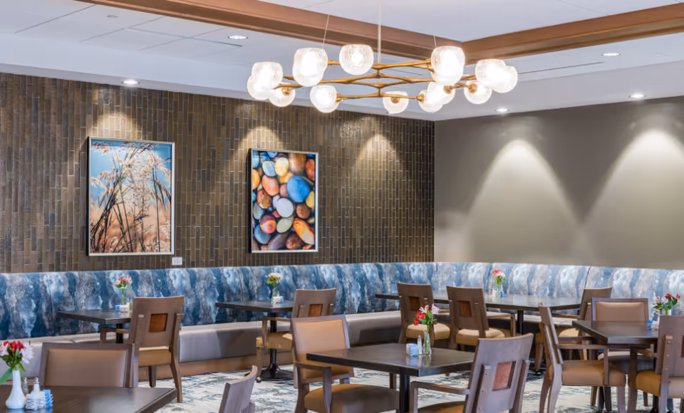 A dining area with multiple wooden tables and chairs, each table decorated with small flower vases. The room features a long blue patterned bench along the wall, two framed pictures on a textured brown wall, and a modern chandelier with multiple round lights hanging from the ceiling.