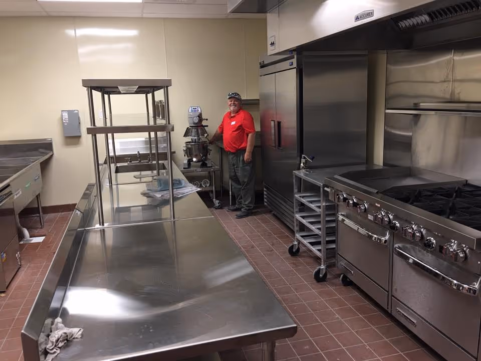 A stainless-steel commercial kitchen with work tables, large stove and refrigerator, and a person in a red shirt standing near a mixer.