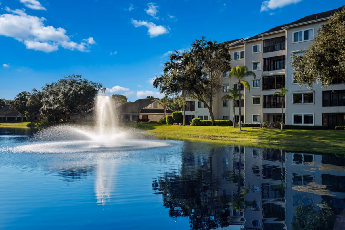 A serene outdoor scene at Arbor Trace featuring a large pond with a water fountain spraying water upwards. The pond reflects the blue sky, scattered clouds, trees, and a multi-story residential building with balconies. The grassy area around the pond is well-maintained with a few palm trees and other greenery.