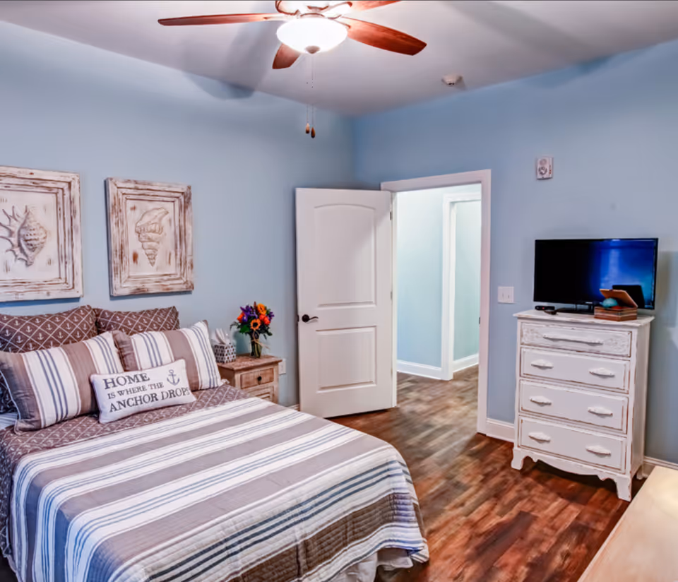 A cozy bedroom with light blue walls, a ceiling fan with wooden blades, and a bed with striped bedding and multiple pillows including one that says 'HOME IS WHERE THE ANCHOR DROPS'. There are two framed seashell artworks on the wall above the bed. A wooden nightstand with a vase of flowers and a tissue box is next to the bed. Across from the bed is a white distressed dresser with a flat-screen TV on top. The door to the room is open, showing a hallway with similar flooring and light blue walls.