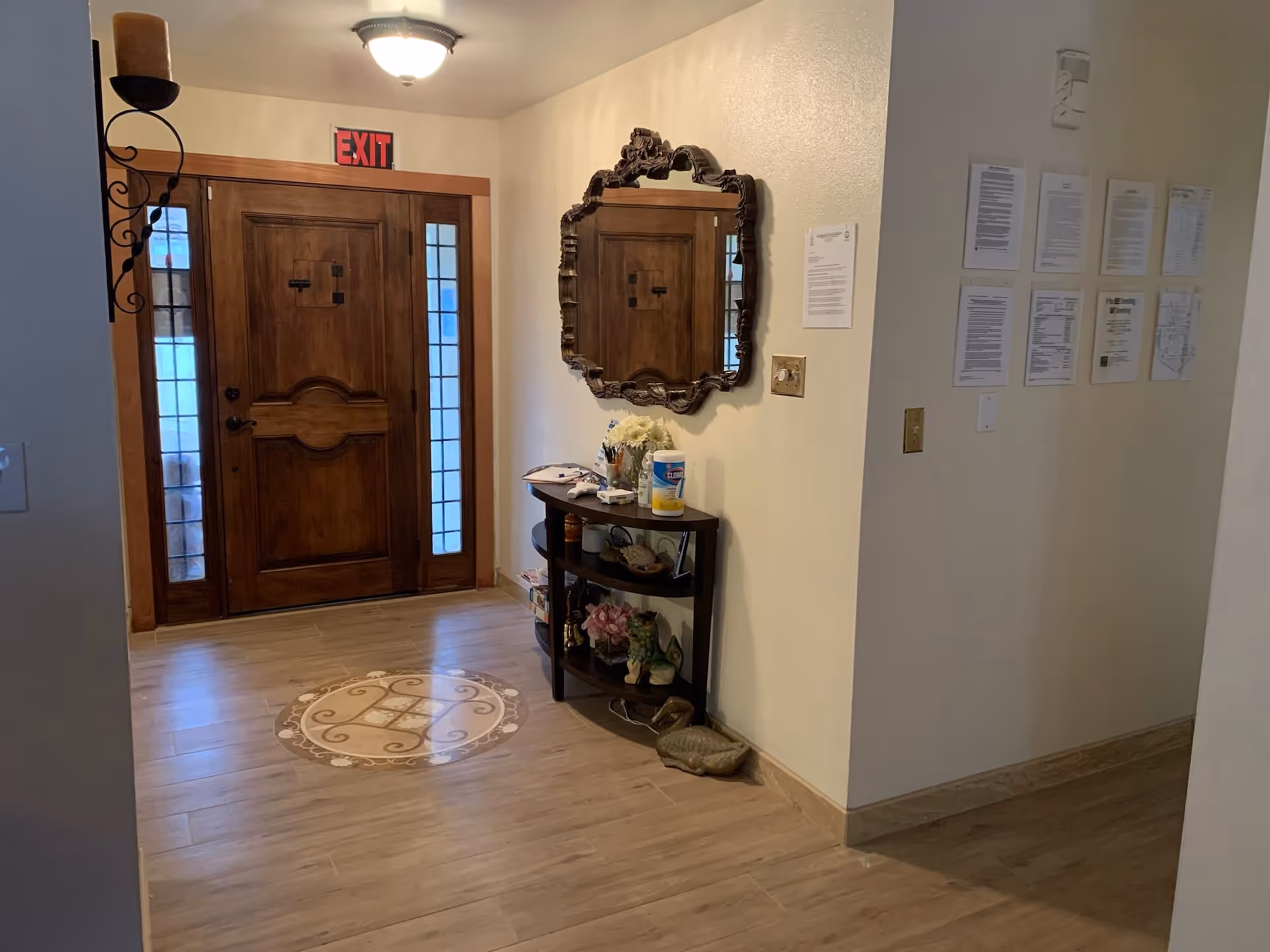 Interior view of an entryway with a wooden front door featuring glass panels on each side. Above the door is a red EXIT sign. To the right of the door is a decorative wall mirror with an ornate frame hanging above a small curved table holding various items including a container of Clorox wipes, flowers, and other small objects. The floor has a circular decorative tile pattern near the door. Several papers and notices are posted on the adjacent wall.